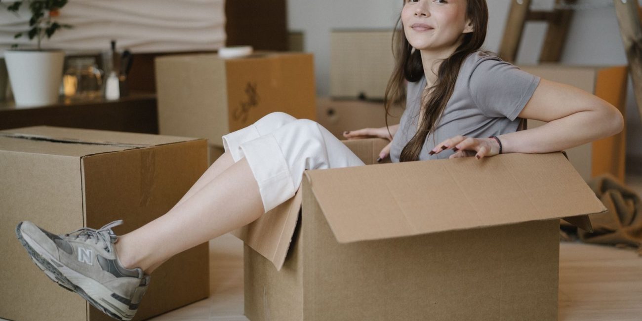 A cheerful young woman sits comfortably in a cardboard box, surrounded by more boxes in a cozy indoor setting.