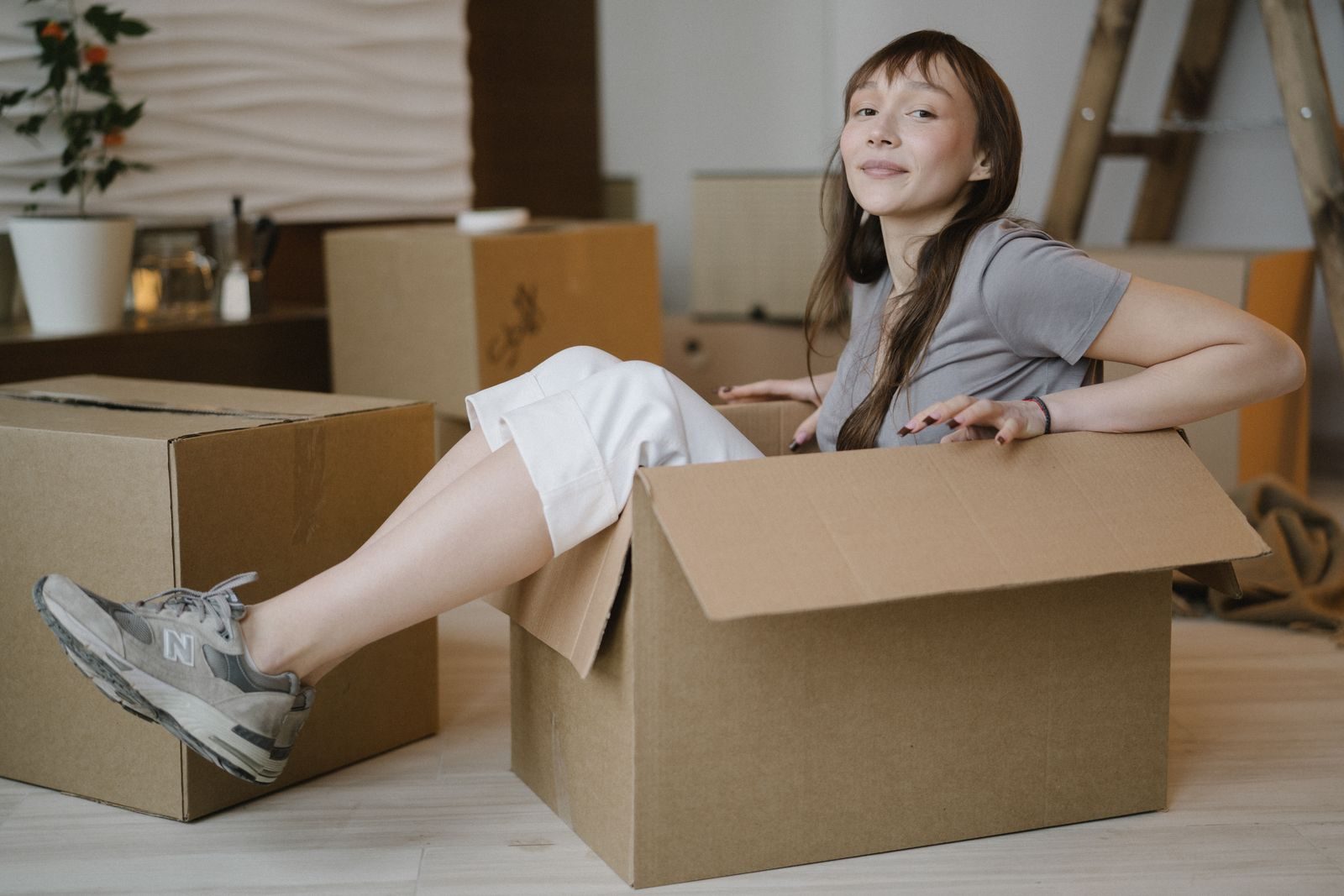A cheerful young woman sits comfortably in a cardboard box, surrounded by more boxes in a cozy indoor setting.