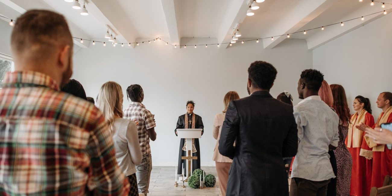 A diverse congregation stands together in prayer led by a woman pastor indoors.